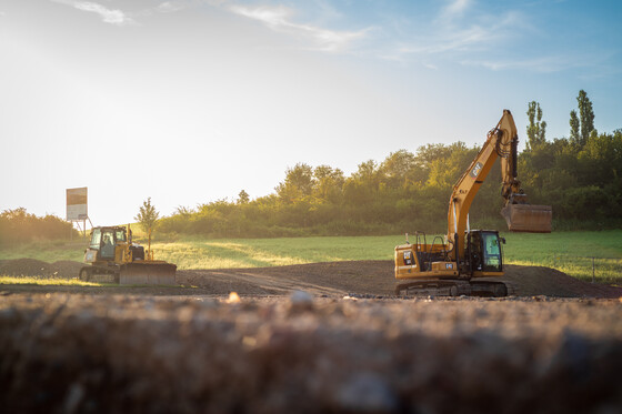 Abendlicht über Baustelle mit mehreren Baggern in leicht hügeliger Landschaft.